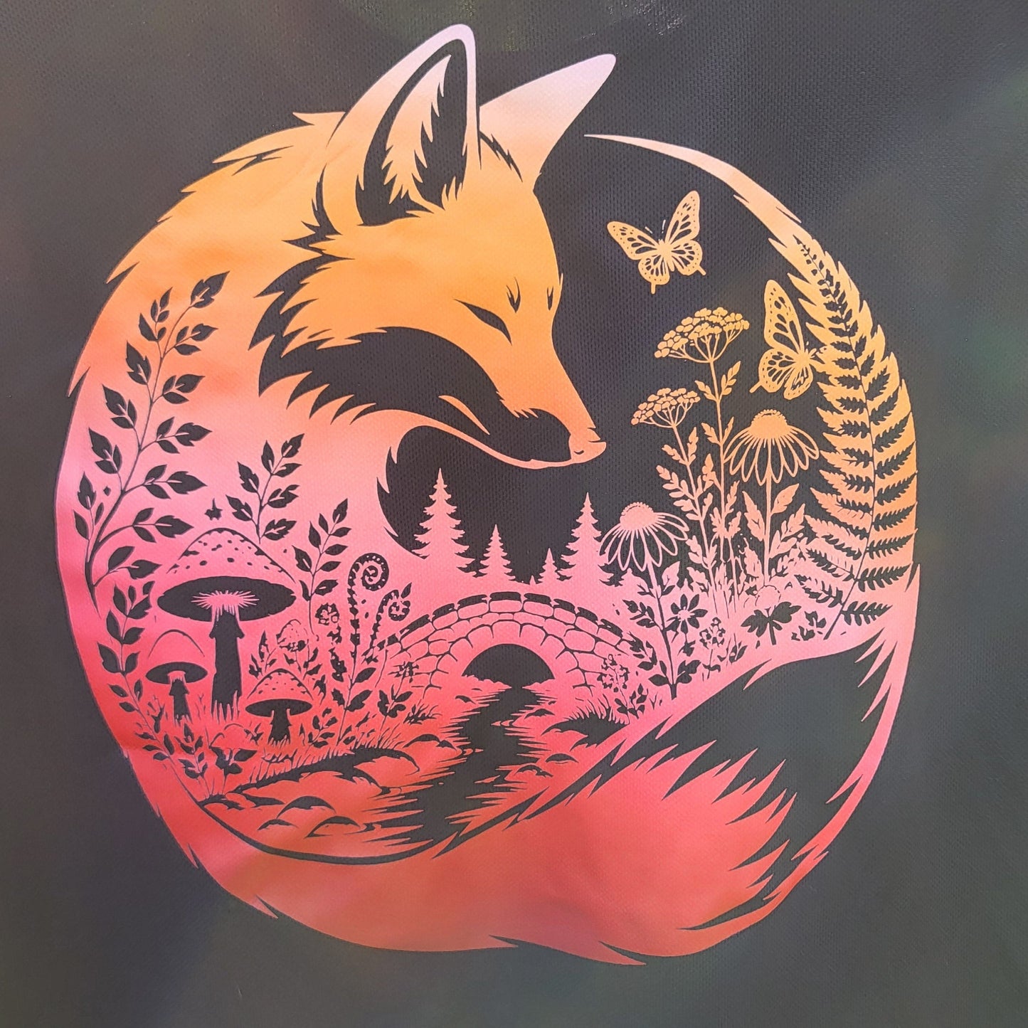 A black t-shirt featuring a large circular graphic of a fox in a forest with a warm orange-to-pink gradient and gold butterfly accents, photographed outdoors on a grassy lawn.
