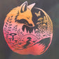 A black t-shirt featuring a large circular graphic of a fox in a forest with a warm orange-to-pink gradient and gold butterfly accents, photographed outdoors on a grassy lawn.