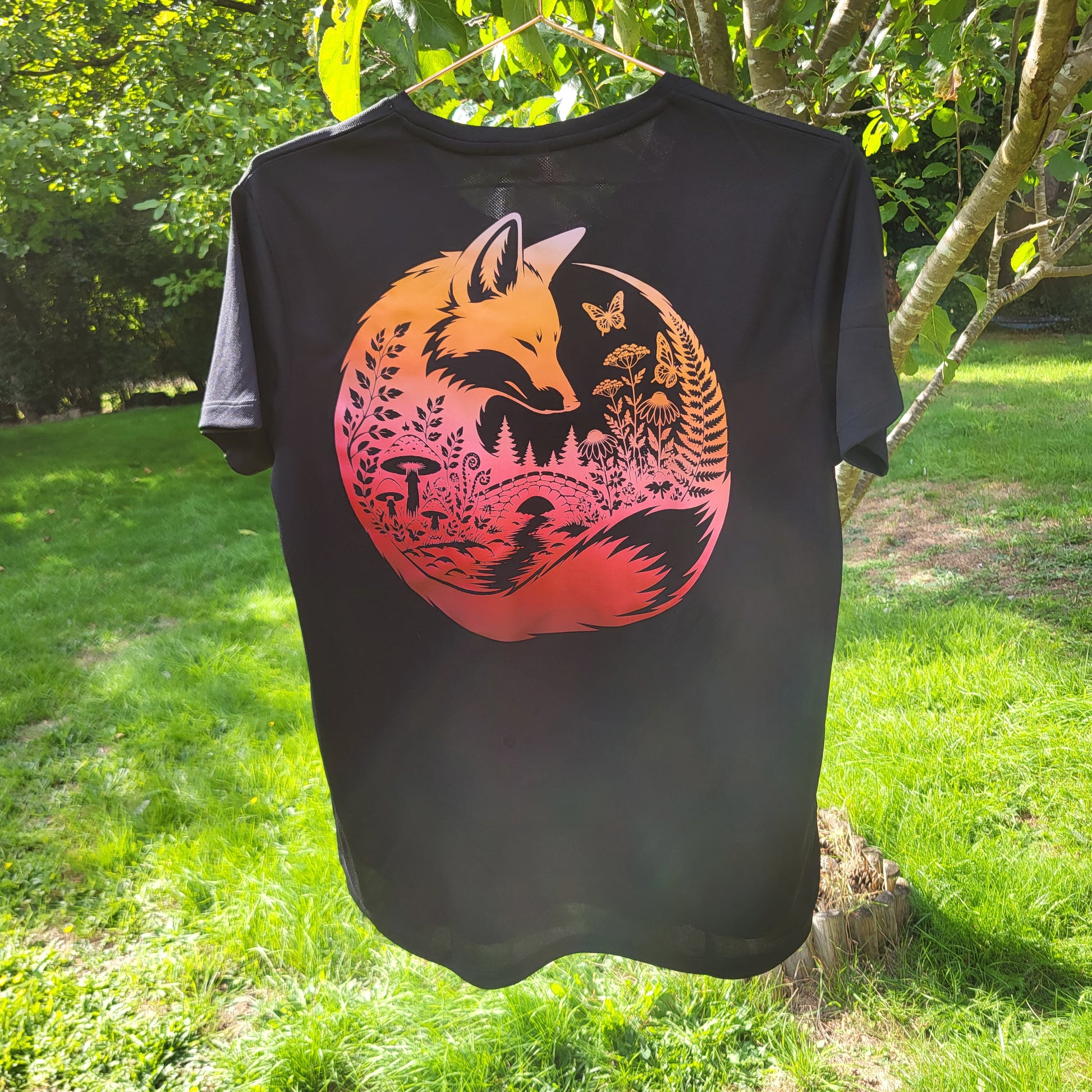 A black short-sleeve t-shirt hanging outdoors on a tree in a grassy garden. The back of the shirt features a large circular graphic of a fox surrounded by forest and nature elements in orange, pink, and red tones.