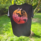 A black short-sleeve t-shirt hanging outdoors on a tree in a grassy garden. The back of the shirt features a large circular graphic of a fox surrounded by forest and nature elements in orange, pink, and red tones.