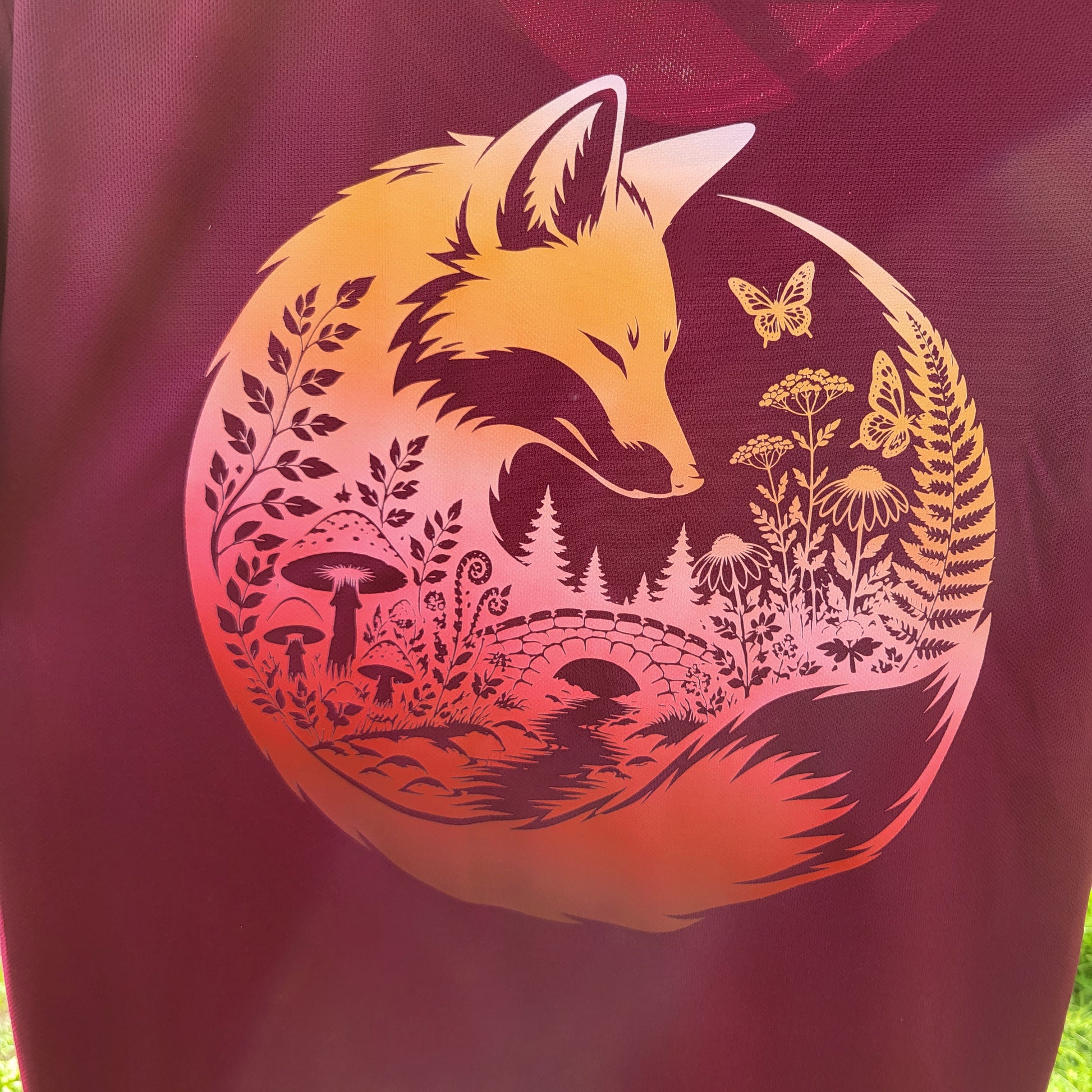 A close-up of a dark burgundy or maroon fabric featuring a large circular graphic print of a fox surrounded by a forest scene in yellow, orange, and pink tones.
