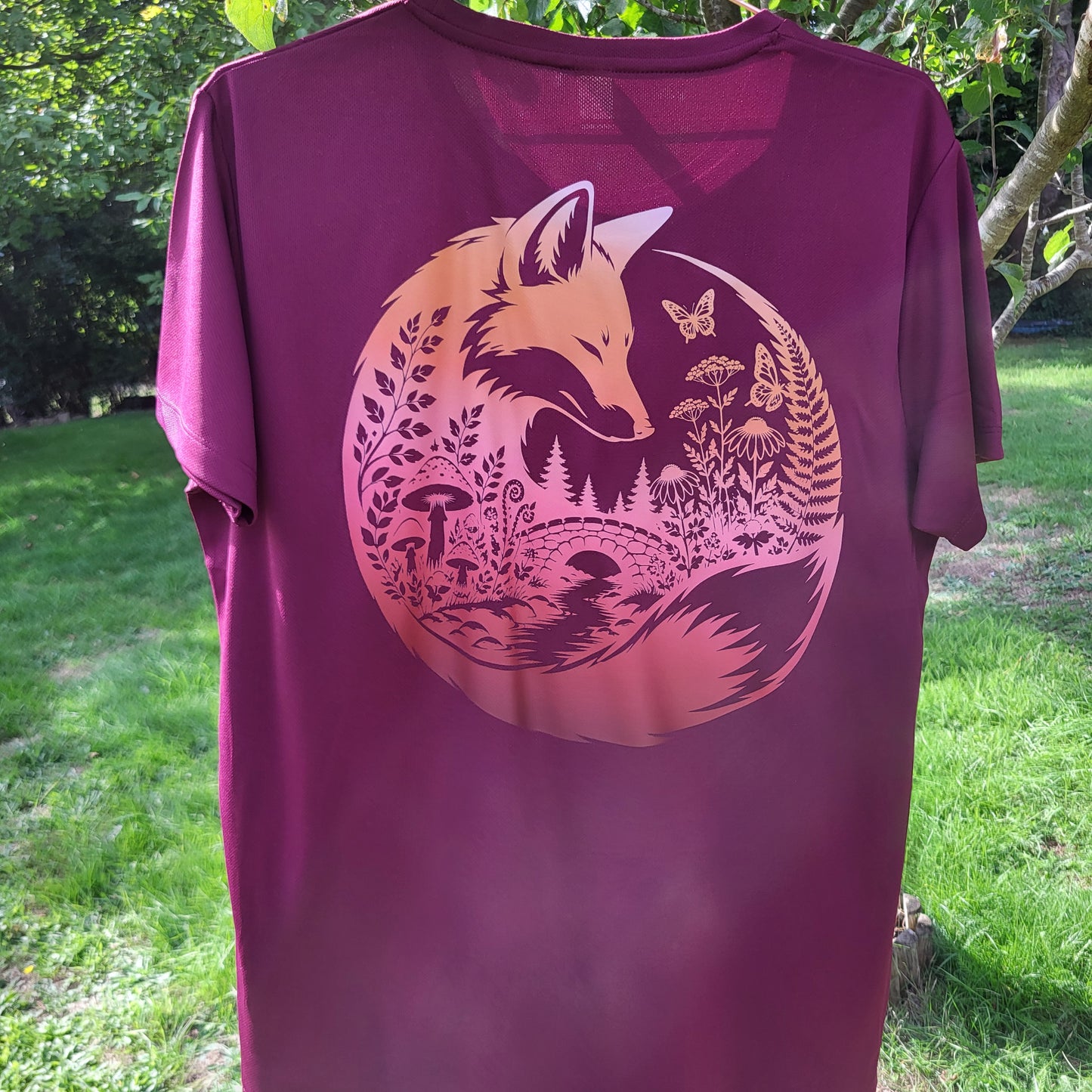 A burgundy short-sleeve t-shirt with a large circular graphic on the back showing a fox, ferns, mushrooms, and butterflies in a pink-to-orange gradient, photographed outdoors on a grassy lawn with trees in the background.