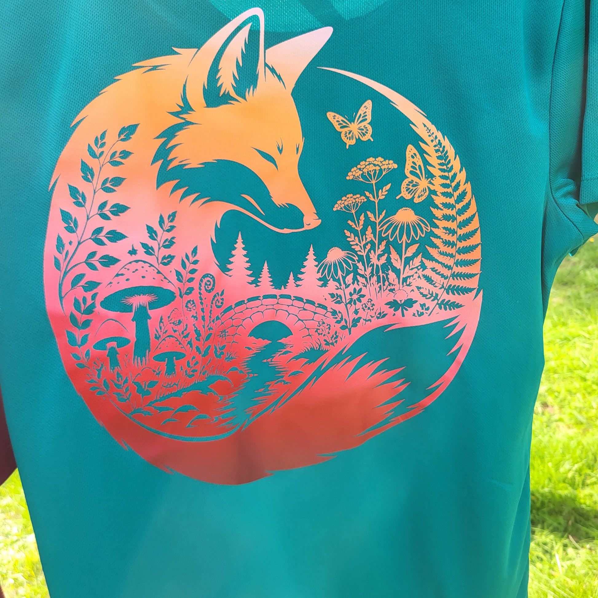 A close-up of a teal t-shirt featuring a large circular graphic of a fox surrounded by nature elements like flowers, mushrooms, and trees, with a sunset gradient transitioning from orange to pink. The shirt is photographed outdoors on grass.