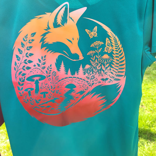 A close-up of a teal t-shirt featuring a large circular graphic of a fox surrounded by nature elements like flowers, mushrooms, and trees, with a sunset gradient transitioning from orange to pink. The shirt is photographed outdoors on grass.