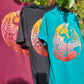 Three short-sleeve crewneck t-shirts displayed outdoors on a hanger against a leafy background. The shirts are in different colors: a teal green shirt in front, a black shirt in the middle, and a burgundy shirt in the back.