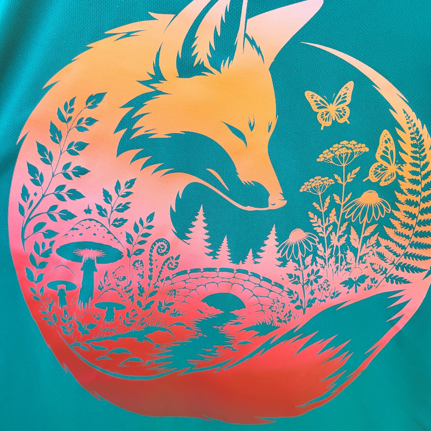A close-up of a circular graphic print featuring a fox head and forest motifs in yellow and orange tones over a teal background.