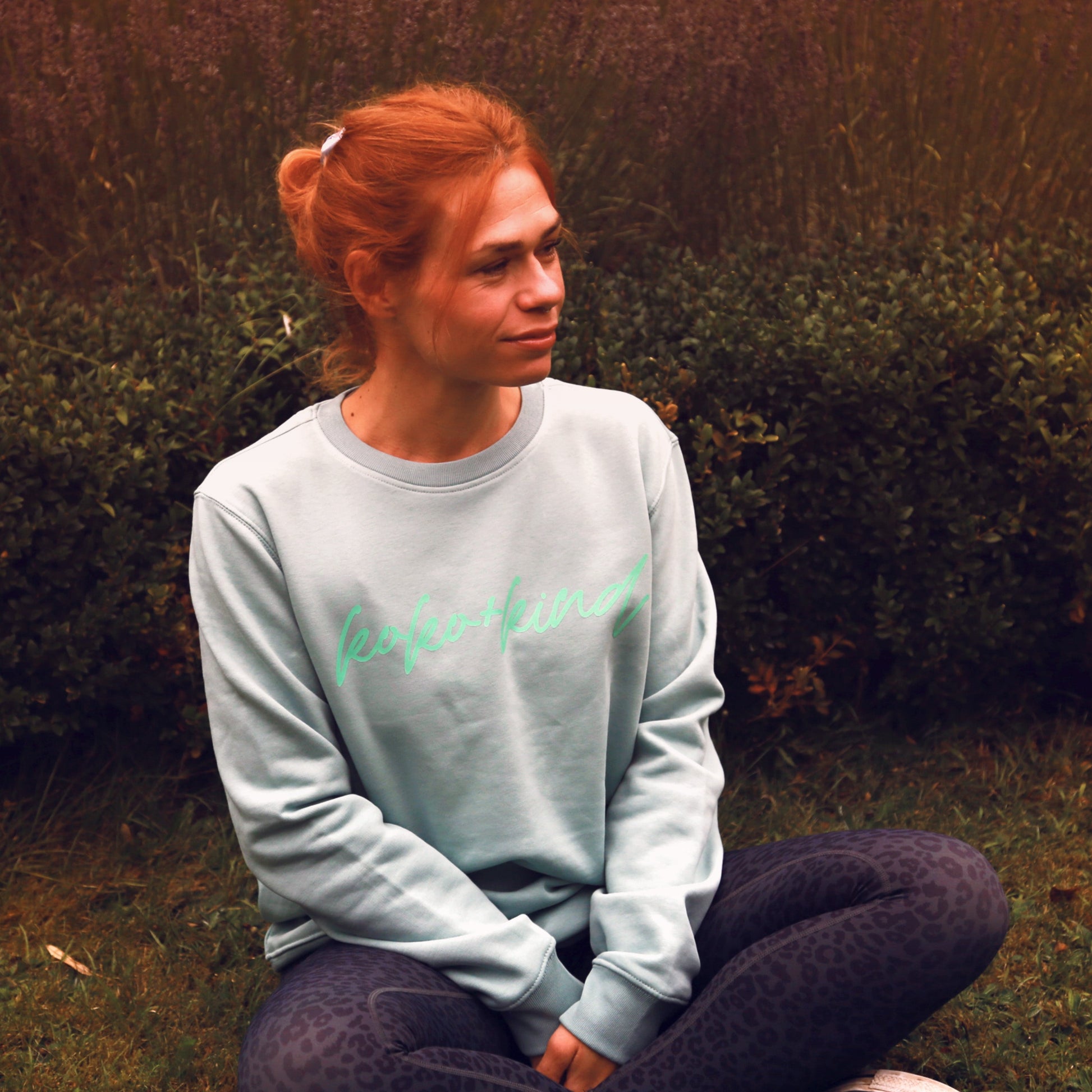 Beccy sits on the floor wearing Sage green Sweatshirt with koko+kind accross the chest also in green. She is also wearing the Shadow Leopard Black Leggings.